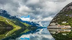 Foto eines Gewässers umgeben von einer Berglandschaft. Am Ufer befindet sich ein längliches Gebäude.