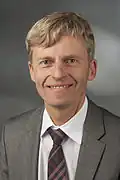 Das Portraitfoto eines mittelalten blonden Mannes, der in die Kamera lächelt. Er hat graue Augen. Er trägt ein weißes Hemd, eine rot-schwarz-karierte Krawatte und ein graues Sakko.