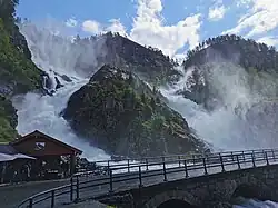 Foto zweier direkt nebeneinanderliegenden Wasserfälle in felsiger Landschaft. Im Vordergrund befindet sich eine steinerne Straßenbrücke.
