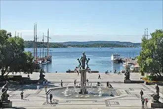 Foto eines direkt am Fjord gelegenen gepflasterten Platzes mit einem Brunnen im Zentrum. Im Brunnen eine größere Statue. Der Brunnen selbst von vier kleineren Statuen umrandet.