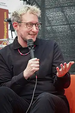 Linus Giese auf einer Couch sitzend mit einem Mikrofon in der Hand