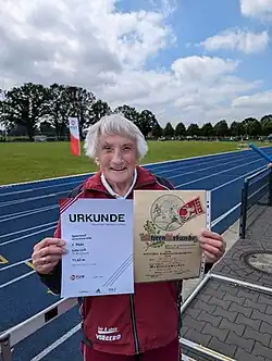 Lotte Leiß steht im Stadion und trägt einen roten Trainingsanzug. Sie zeigt zwei Urkunden von Westfalenmeisterschaften: die eine von 2024, die andere von 1954.