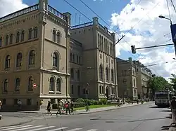 Seitliche Farbfotografie von einem grauen, dreigeschossigen Gebäude mit drei Vorbauten, die an den Ecken und in der Mitte sind. Leichte Ornamente sind an den Ecken und über den Rundbogenfenstern. Auf den Dächern sind Steingeländer, die von Eckpfeilern gestützt werden.