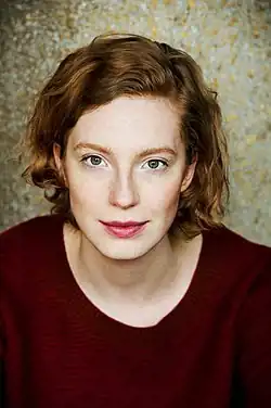 Porträt von Luise Wolfram, 2017 mit lockigem, hellbraunem Haar und trägt einen dunkelroten Pullover. Ihr Gesichtsausdruck ist freundlich. Der Hintergrund besteht aus einer strukturierten Oberfläche mit einer Mischung aus goldenen und grauen Farbtönen.