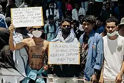 Demonstranten in Osttimor mit Schildern. Links: „Ich bin hässlich wegen der Tattoos, aber ich bin kein Mafioso wie die 65 Abgeordneten“. Rechts: „Beendet die lebenslangen Pensionen, nieder mit den geizigen Abgeordneten.“