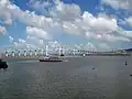 Die Brücke ist aus der Ferne zu sehen. Im Vordergrund ist Wasser und ein Boot. Der Himmel ist blau und leicht wolkig.