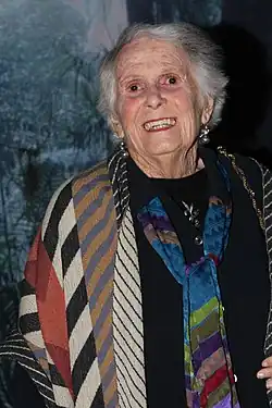 Margaret Fulton steht vor einer Glasscheibe, hinter der sich mehrere Palmen befinden. Sie trägt ein schwarzes Oberteil mit einem schwarz-gold geflochtenen Schmuckstück auf der linken Schulter, einen mehrfarbig gestreiften Blazer, eine breite, mehrfarbig gestreife Krawatte, über der Krawatte eine silberne Olivenzweig-Brosche mit zwei grünen Steinen und silberne Kugelohrringe. Ihre weiße, flach abstehende Frisur trägt sie bis zum Nacken.