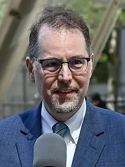 Nahaufnahme eines mittelalten weißen Mannes hinter einem Mikrofon, unscharfer Hintergrund mit einem Gebäude und Straßenmöblierung. Dunkles, schütteres Haupthaar, dunkler und weißer, kurzer Oberlippen- und Kinnbart, Brille mit dunklem Gestell. Business-Kleidung mit blauem Jackett, weißem Hemd, grauer Krawatte.