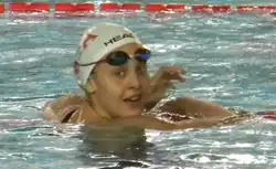 Carraro ist nach einem Rennen noch im Wasser und stützt sich auf die Bahnabgrenzung. Sie trägt eine weiße Bademütze, die Schwimmbrille hat sie auf die Stirm hochgeschoben.