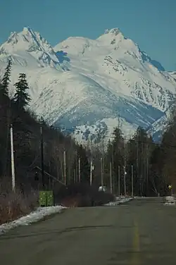 Ein mehrgipfliger Berg ragt über Bäumen und einer asphaltierten Straße auf