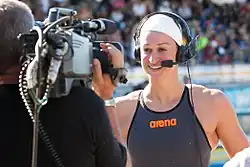 Megan Romano im dunklen Badeanzug mit Weißer Badekappe und darüber einem Headset wird gerade interviewt. Die linke Bildhälfte zeigt den Kameramann von hinten.