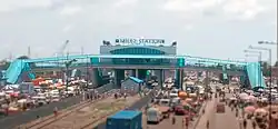 Panoramafoto: Frontal das Stationsgebäude, erhöht über dem Expressway, der vom Bildhintergrund in den Vordergrund verläuft. Viele Autos auf dem Expressway.