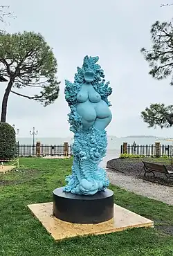 vor dem Hintergrund des Canale grande in Venedig steht eine türkisgrün gefasste Statue. Eine abstrahierte weibliche Figur in Rückansicht mit Brüsten am Rücken, umgeben von Ornamenten und Metamorphosen zwischen Frau, Fisch, Pflanze und Ornament. Eine heutige Fruchtbarkeitsgöttin.