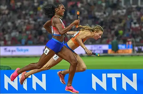 Alexis Holmes in rot-weiß mit Staffelstab in der Hand zieht an Femke Bol in Orange, die stolpert und die Kontrolle über den Staffelstab verliert, vorbei zu WM-Gold.