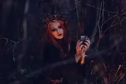 Eine Wicca sitzt auf dem Waldboden. Sie trägt eine Krone aus Ästen und hält einen Kelch.