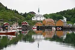 Foto einer weißen Holzkirche. Im Vordergrund ein Gewässer mit mehreren Booten, an dessen Ufern kleinere Holzgebäude stehen.