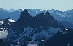 Ein dunkler zerklüfteter Berg ragt über Gletschereis im Vordergrund und vergletscherte Berge im Hintergrund.