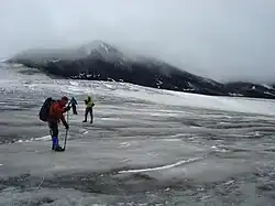Drei Bergsteiger verlassen einen Gletscher an einem wolkigen Tag; im Hintergrund ein felsiger Gipfel.