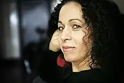 Nahid Persson Sarvestani schaut seitlich in die Kamera. Das Farbfoto zeigt sie im Porträt. Sie hat langes lockiges schwarzes Haar und trägt dunkle Kleidung.