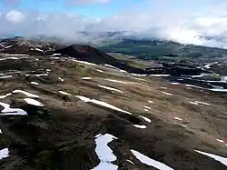 Ein dunkel gefärbter Vulkankegel mit einem Gipfelkrater ragt über einer spärlich mit Schnee bedeckten vegetationsarmen Hochebene auf.