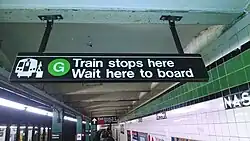 U-Bahn-Station, Blechschild hängt quer über Bahnsteig, Beschriftung in weiß auf schwarz: „G / Train stops here / Wait here to board“ nebst stilisierter Darstellung eines Zugs mit Bahnsteig und einsteigender Person