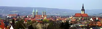 Von erhöhter Position aufgenommen, rechts ist über den Häusern der Stadt den Turm der Stadtkirche zu sehen, links sieht man die vier Türme des Doms. Im Hintergrund ist der Hang des Saaletals in größerer Entfernung zu sehen.