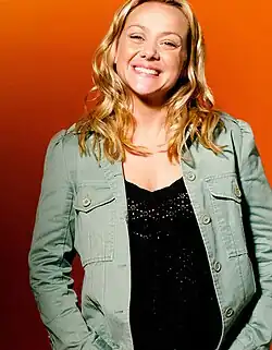 Nicole Sullivan mit schulterlange blonde, wellige Haare im Jahr 2003 vor einem orangefarbenen Hintergrund, lächelt breit. Sie trägt eine hellgrüne Jacke mit Knöpfen und Taschen, die sie über einem schwarzen Oberteil mit funkelndem Design trägt.