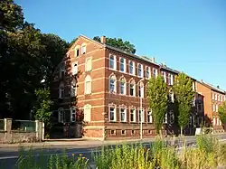 zwei vierstöckige Häuser, dreispurige Straße (davon zwei Spuren mit Pfeilmarkierungen zum Abbiegen), Grünbewuchs