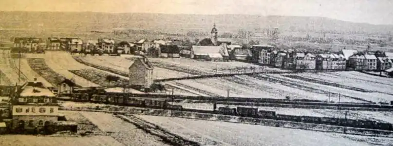 Oberndorf in den 1910er Jahren, im Hintergrund der Steigerwald. Die Bahnstrecke nach Erfurt (vorne) bildet die Grenze zwischen den heutige Stadtteilen Oberndorf und Bergl. Die Bahnstrecke unmittelbar dahinter führt nach Würzburg. Vorne links das Oberndorfer Wasserhaus von 1911 und am rechten Ortsrand der Häuserblock Kornstraße/Bismarckstraße (heute Engelbert-Fries-Straße). Unmittelbar links des noch bestehenden Hauses links der Bildmitte verläuft heute die Franz-Josef-Strauß-Brücke (Bahnüberführung).