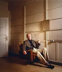 Richard Ford sitzt auf einer Couch und hält die Faust hoch, hinter ihm eine eingeschlagene Wand.