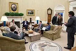 In einer Sitzung mit gemütlicher Atmosphäre ist Barack Obama in Gespräch mit Beratern. Die Teilnehmer sitzen auf Sofas und Sessel rund um einen niedrigen Fliesentisch mit einer Obstschale darauf