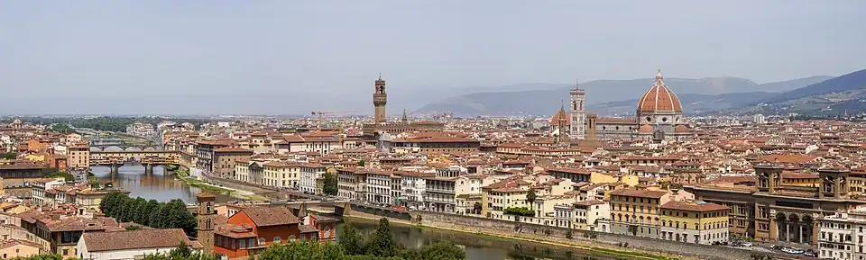Farbiges Stadtpanorama mit einem Fluss, der von unten rechts bis zur linken Mitte führt. Mehrere alte Bogenbrücken verbinden beide Ufer und eine Brücke hat einen überdachten Gang. Rechts ist ein großer Kuppelbau und in der Mitte ein rechteckiger Turm. Im Vordergrund steht ein Park mit Hecken und Palmen und im Hintergrund sind Berge.