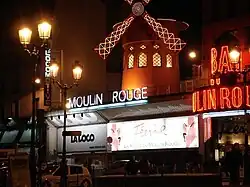 Die Frontseite der Gebäude mit dem Moulin Rouge, mit markanter Windmühle und Neonbeleuchtung, sowie das Schild La Loca für den Club La Locomotive.