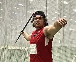 Mann im mittleren Alter im roten Sporttrikot und schwarzen Kopfschweißband im Seitenprofil und in die Kamera schauend mit ausgestreckten Armen und in der linken Hand einen Speer haltend in einem gitterartigen halboffenen Schutzzelt.