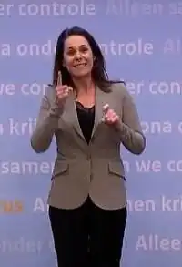 Gebärdendolmetscherin Irma Sluis, die vor einem Hintergrund mit Text steht, trägt einen hellen Blazer über einem dunklen Oberteil und dunklen Hosen. Sie scheint gerade durch Handgesten, das gesprochene Wort in Gebärdensprache auszudrücken. Der Hintergrund enthält wiederholten Text in mehreren Sprachen, darunter: „we control“, „Alleen samen“, „alleen controle“, „samen we“ Auf der linken Seite des Hintergrunds ist das Wort „virus“ in orange Farbe zu sehen.