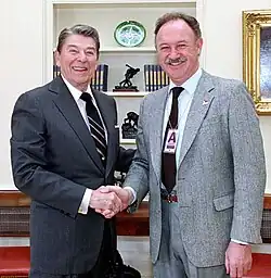 Gene Hackmann im grauen Anzug mit hellblauem Hemd und schwarzer Krawatte. Um seinem Hals ein Ausweisanhänger in einer Kunststoff Schutzhülle mit dem Buchstaben A an einer silbernen Kette. Schüttelt die rechts Hand des US-Präsidenten Ronald Reagan, ebenfalls einen Anzug und Krawatte tragend im Oval-Office am 6. Februar 1987 aufgenommen. Im Hintergrund ein Bild in einem goldenen geschwungenen Rahmen und einem Bücherregal mit 3 Regalböden bestückt mittig einem Teller mit einem Symbol, darunter eine Statue umrahmt von Büchern.