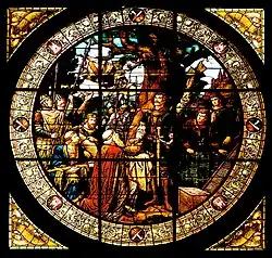 Rundfenster mit farbigem Glasbild, das eine mittelalterliche Szene mit mehreren Personen um einen Baum zeigt, darunter ein Ritter und weitere Figuren in historischer Kleidung