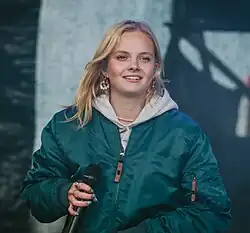 Paula Hartmann lächelt, während ihres Auftritts beim Rocken am Brocken 2022. Sie trägt eine grüne Bomberjacke über einem grauen Kapuzenpullover. In ihrer rechten Hand hält sie ein Mikrofon, während sie scheinbar inmitten eines Songs oder einer Ansprache ist. Ihre blonden Haare fallen locker um ihr Gesicht, und sie trägt auffällige Ohrringe. Der Hintergrund ist verschwommen, aber eine Bühne und Bühnenbeleuchtung sind erkennbar, was die Konzertatmosphäre unterstreicht.