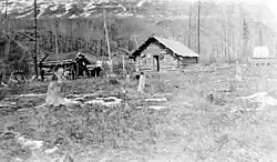 Schwarzweiß-Foto zweier Holzhütten in einem lichten Wald