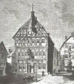 Um 1865: Mit Doppelportal, Rundbogenfenstern im EG und Baldachin überm Balkon[6]