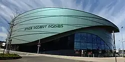 Rundbau, bei dem die Außenverkleidung für den gläsernen Eingangsbereich nach oben geschwungen ist. Auf der Außenverkleidung ist der Schriftzug Stade Robert Poirier in Großbuchstaben angebracht.