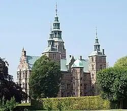 Seitliche Farbfotografie eines Schlosses mit zwei kleinen Dachlaternen auf Seitentürmen und einer großen Dachlaterne in der Dachmitte. Ein Dachgiebel und eine Dachhaube befinden sich zwischen den beiden kleinen Türmen. Auf der Frontseite sind ein Staffelgiebel und zwei vertikale Fenstervorbauten. Im Vordergrund stehen Hecken und Bäume.