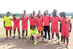 Ein Fußballteam posiert auf einer trockenen Rasenfläche für ein Gruppenfoto. Es sind insgesamt 15 Männer in zwei Reihen stehend, mit dem Torwart vor ihnen hockend. Die erste Reihe hat ihre Arme jeweils über die Schultern der danebenstehenden Teammitglieder gelegt und steht teils leicht nach vorne gebeugt. Im Hintergrund ist ein Waldrand erkennbar.