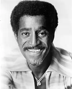 Schwarz-weiß-Porträt eines lächelnden Sammy Davis Junior. Er trägt eine gestreifte Strickjacke und einen Anhänger mit dem Peace-Zeichen um den Hals. Er hat einen schwarzen Schnurrbart und einen kurzen Kinnbart, zudem eine Afrofrisur.