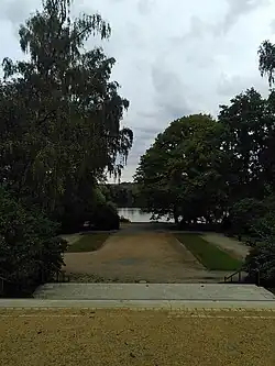 vorne Schotter, dahinter abwärtsführende Treppe, im Mittelgrund breiter Weg mit länglichen Rasenstreifen rechts und links, große Bäume rechts und links ragen in das Bild, hinten der Schwanenteich und das gegenüberliegende bewachsene Ufer