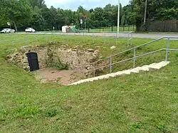 Innerhalb einer Wiese führt eine Treppe abwärts, wo sich eine verschlossene Metalltür befindet. Die Außenwand des Gemäuers ist teilweise sichtbar.