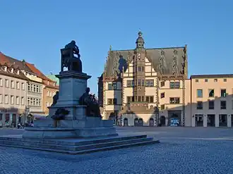 Im Vordergrund eine im Schatten liegende auf einem steinernen Podest stehende Statue. Am Podest sind zwei weitere Statuen platziert. Dahinter steht neben anderen, von der Sonne beschienen Häusern zentral ein mit Wappen verziertes fachwerkartiges Gebäude mit einem zentralen Turm und einer Uhr.