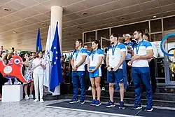 Die vier Personen tragen ein weißes Trikot mit blauer Schulterpartie und blaue Hosen. Um den Hals ist jeweils an einem blauen Band die Medaille erkennbar.