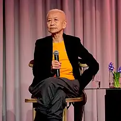 Shu Lea Cheang auf einer Bühne, Mikro in der Hand, neben sich ein Glas Wasser und eine blaue Hyazinthe