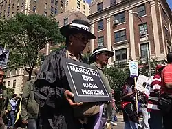 Demonstration. Im Bildvordergrund eine schwarze Frau mit gesenkten Blick. Sie trägt mit beiden Händen ein schwarzes Schild mit der Aufschrift: „MARCH TO END RACIAL PROFILING“
